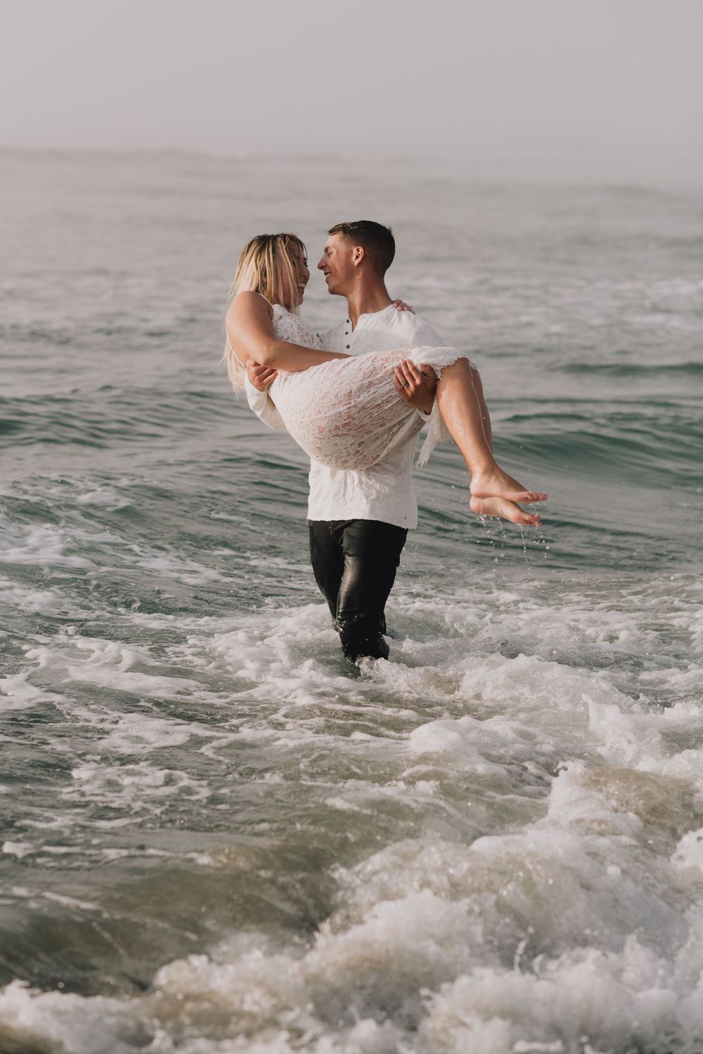 A romantic couple embraces in ocean waves, man carrying woman in white dress through surf at the beach.
