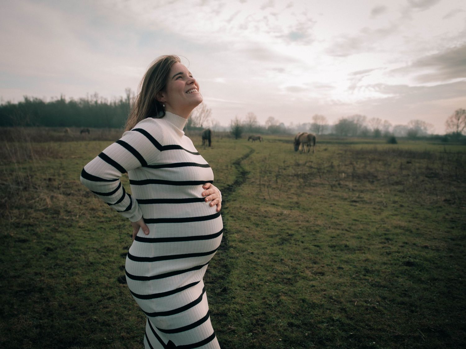 Pregnant woman in striped dress standing in a green field at dusk, holding her baby bump with horses in the background.