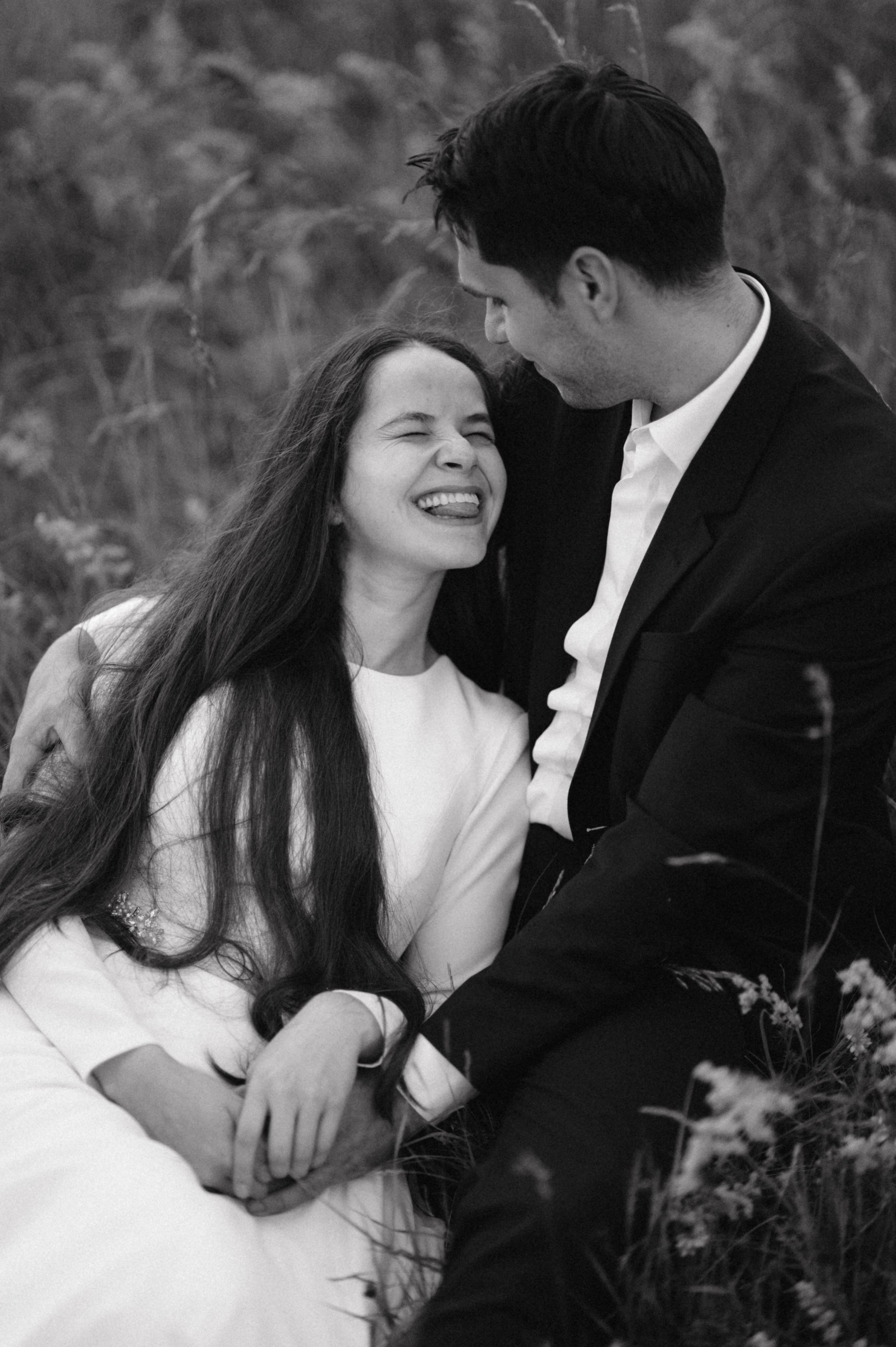 Black and white portrait of couple sharing joyful laughter in a field of tall grass.