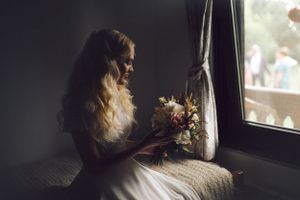 A bride in white dress sits by window holding bouquet in moody lighting.