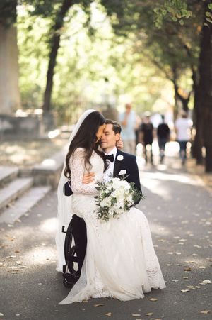 A tender wedding moment between couple on tree-lined path with white bouquet.