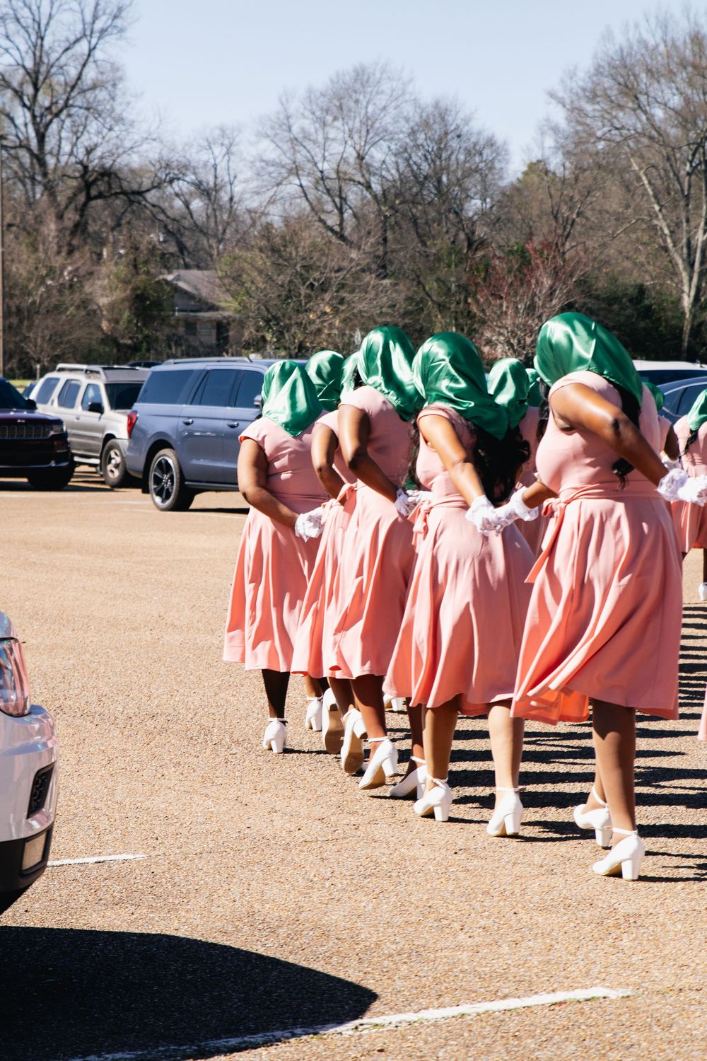 Line of people in matching pink dresses and green headwear standing outdoors on sunny day.
