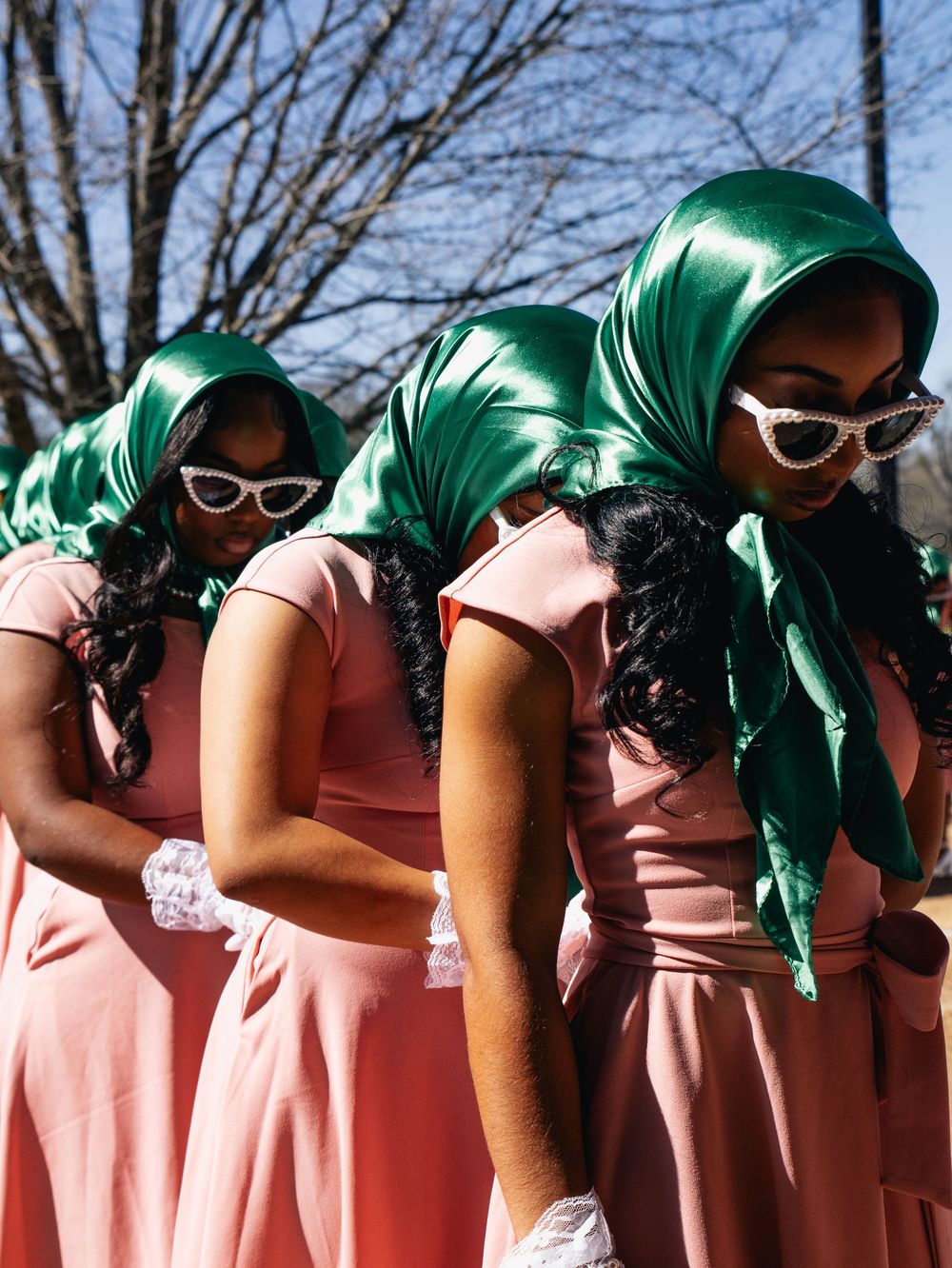 A group wearing matching pink dresses and green headscarves with white sunglasses pose together outdoors.