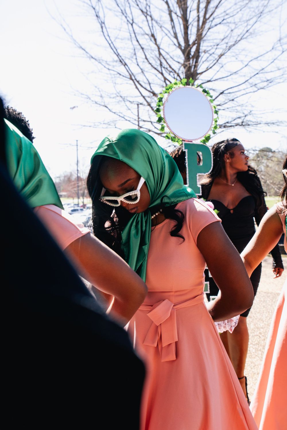 A person in a pink dress and green headscarf with white sunglasses stands among others at an outdoor event.