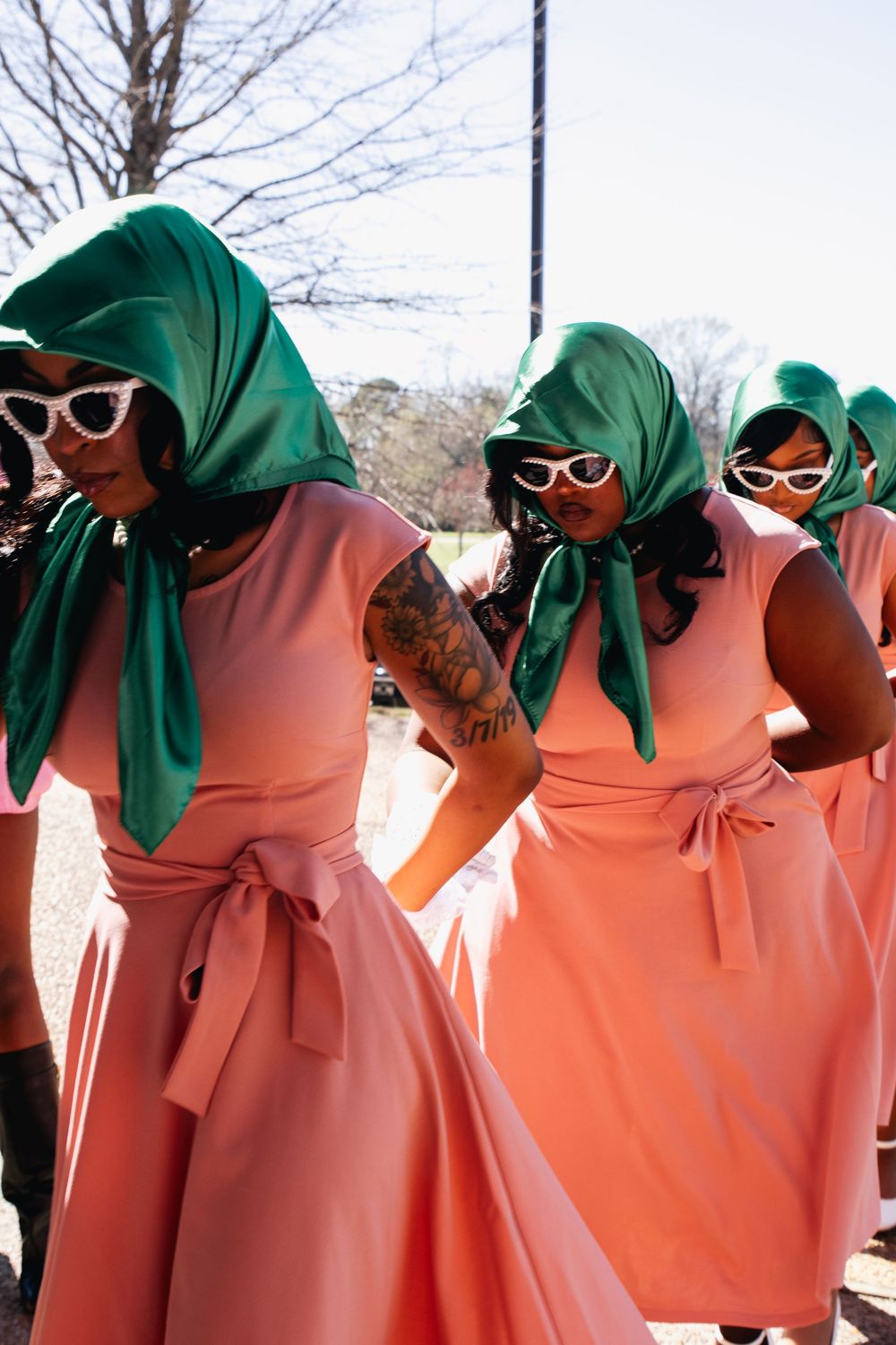 People in coordinated pink dresses and green head coverings with white sunglasses pose together.