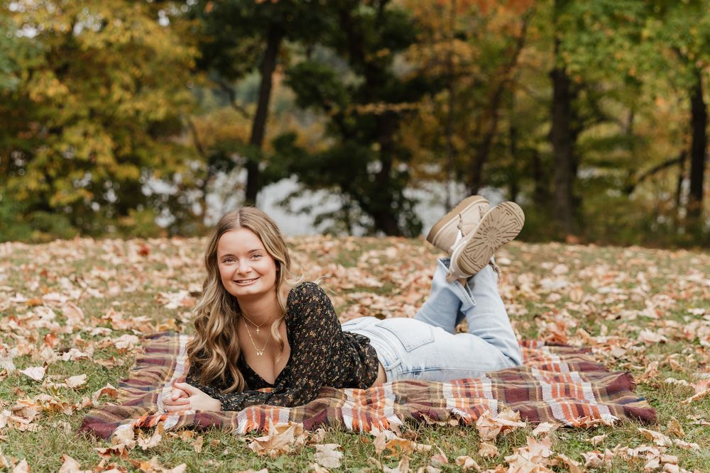 Someone relaxes on a plaid blanket surrounded by fallen autumn leaves in a wooded setting.