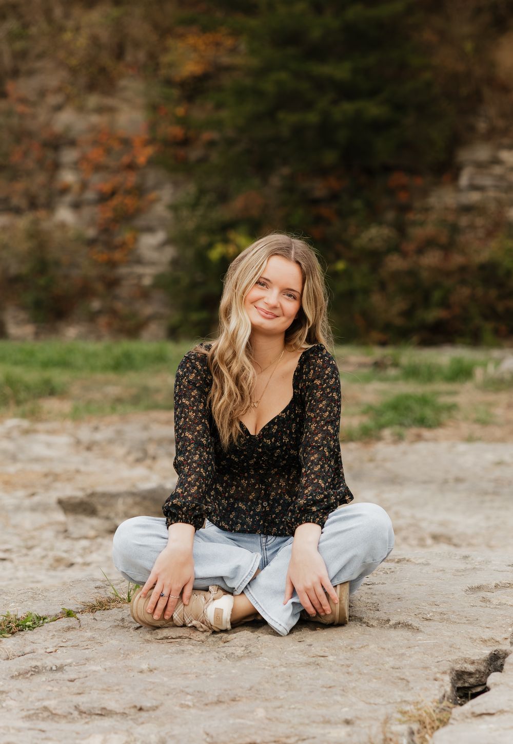 Person in casual attire relaxing on stone surface with fall colors in background.
