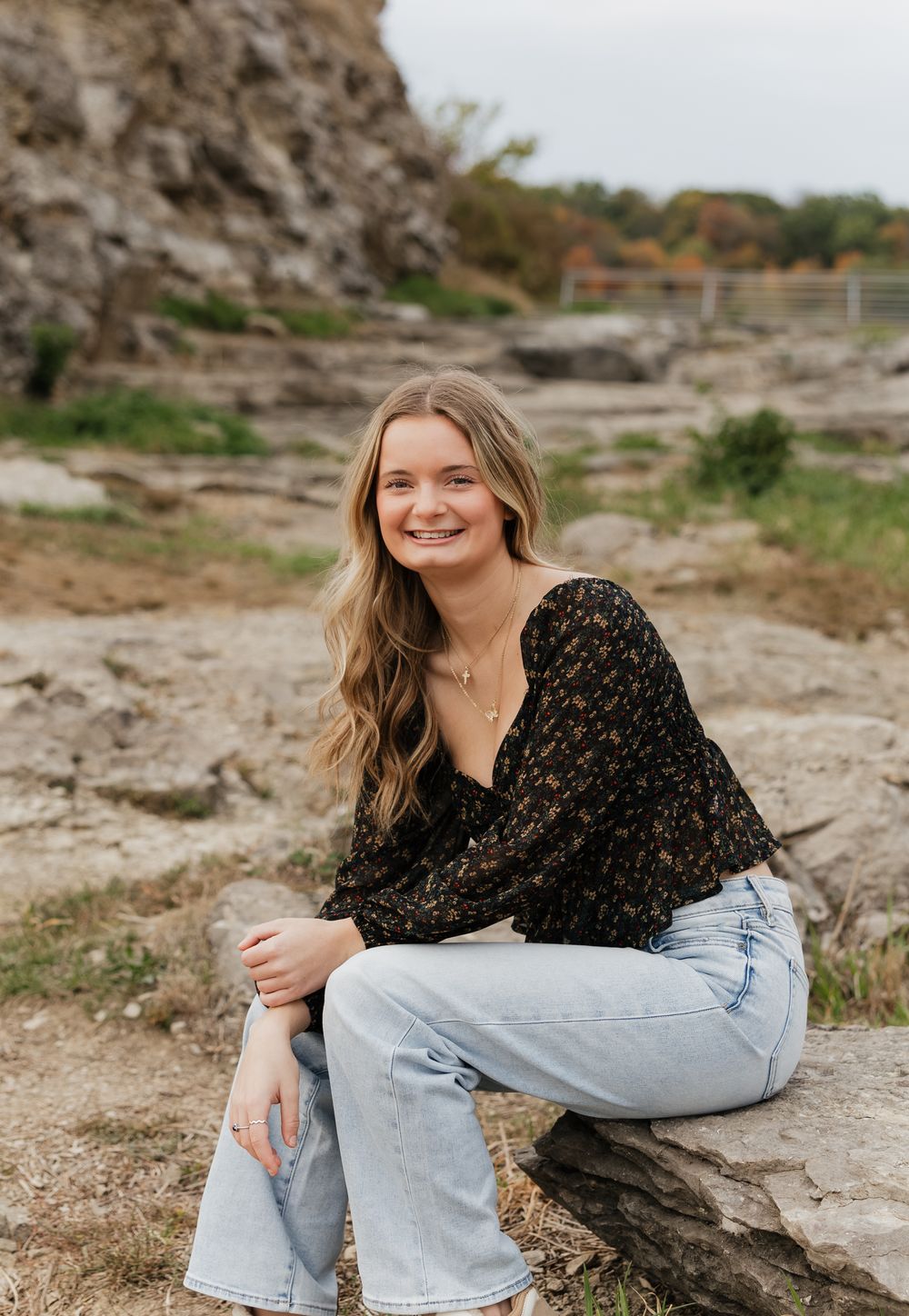 A person in a black floral top and light jeans sits casually on rocky terrain on a cloudy day.