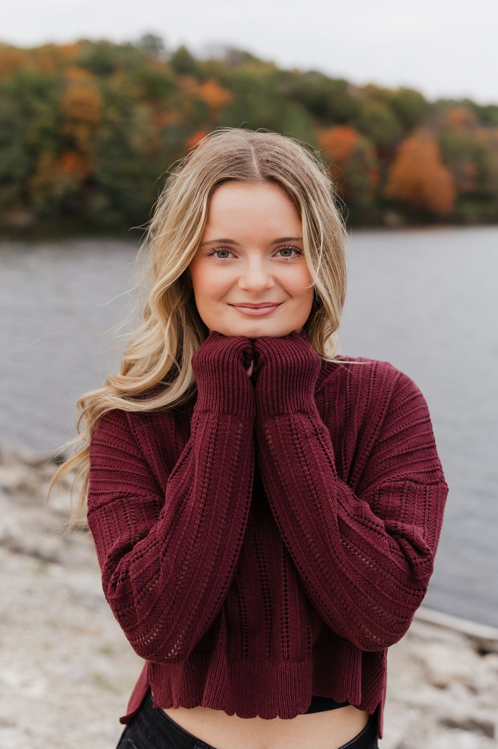 Person in burgundy knit sweater poses by autumn lake with colorful fall foliage in background.