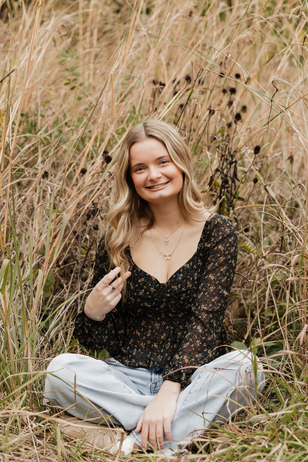 A person wearing a black polka dot top and light jeans poses in tall autumn grass during an outdoor photo session.