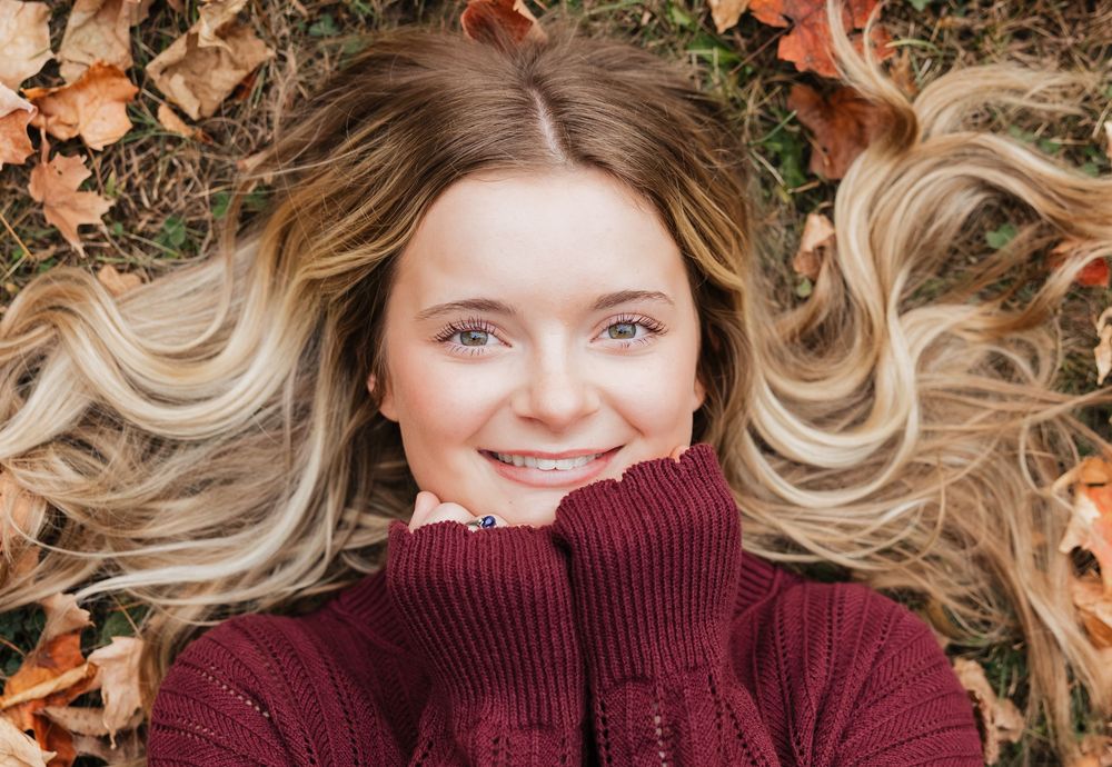 Someone with blonde wavy hair lies on autumn leaves wearing a burgundy turtleneck sweater.