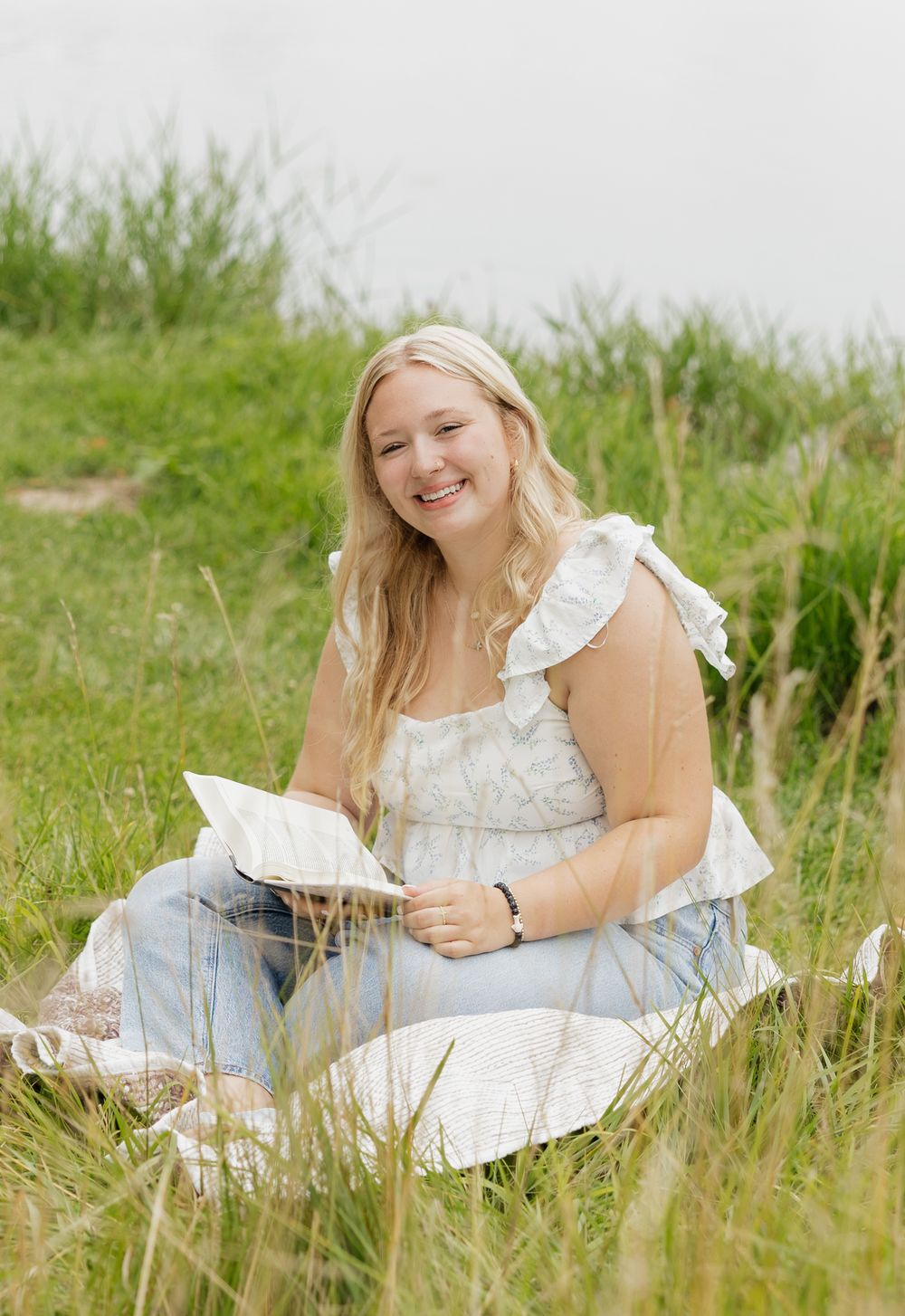 Someone sits peacefully in a grassy meadow wearing a white dress and jeans while reading a book on a sunny day.