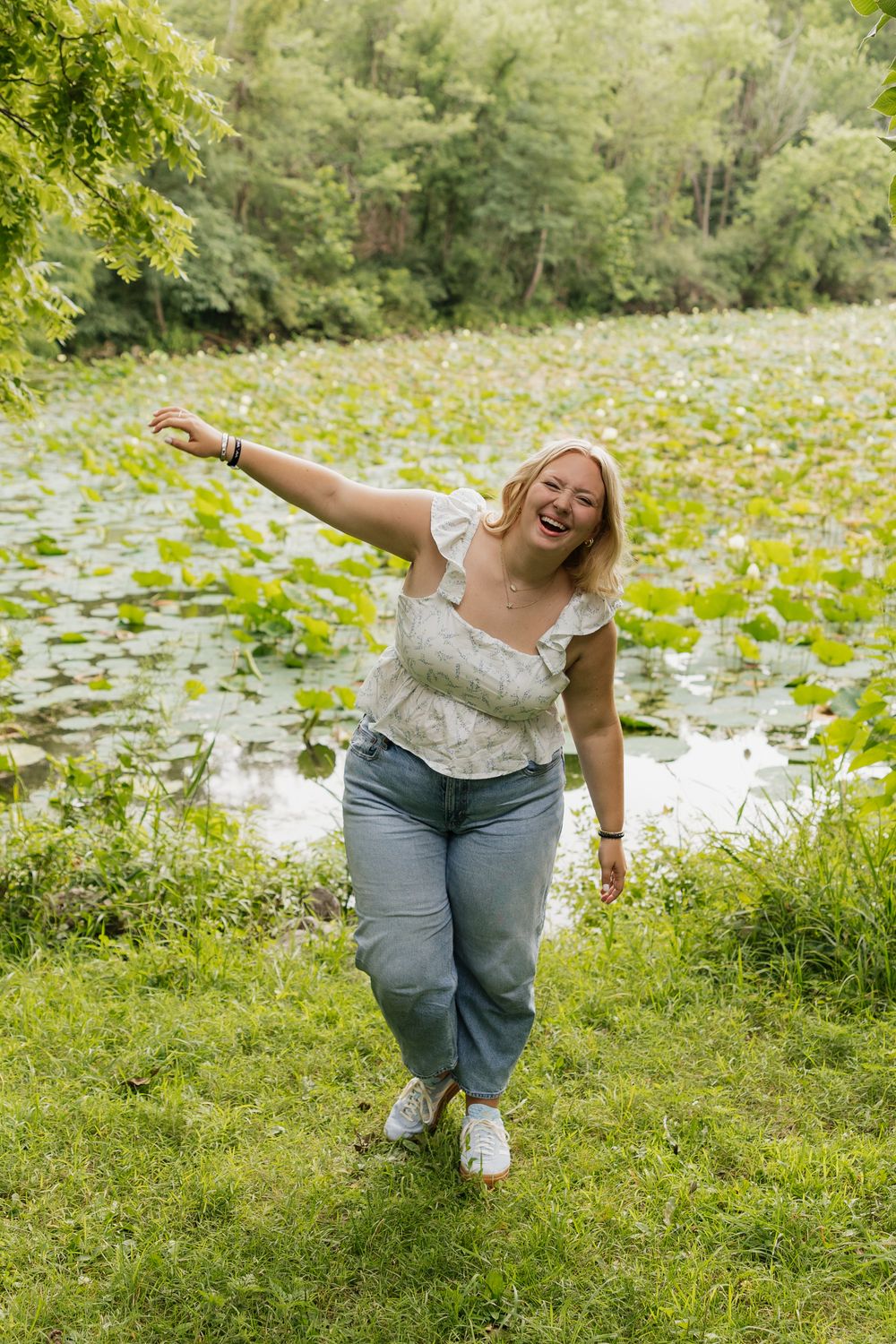 A person in a white top and jeans stands joyfully by a lily pond on a sunny summer day.
