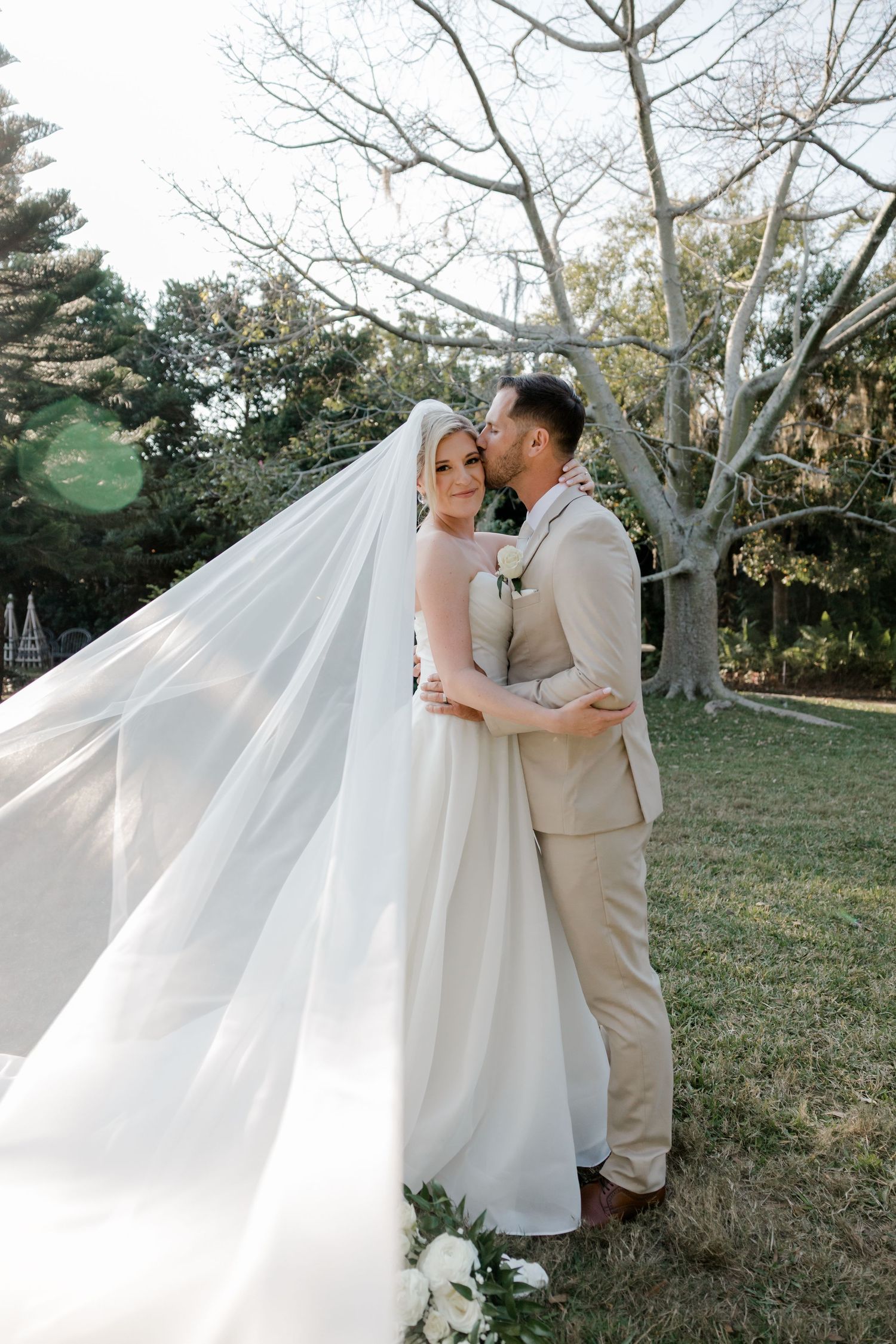 A romantic outdoor wedding photoshoot featuring a couple in white and beige attire embracing under bare winter trees.