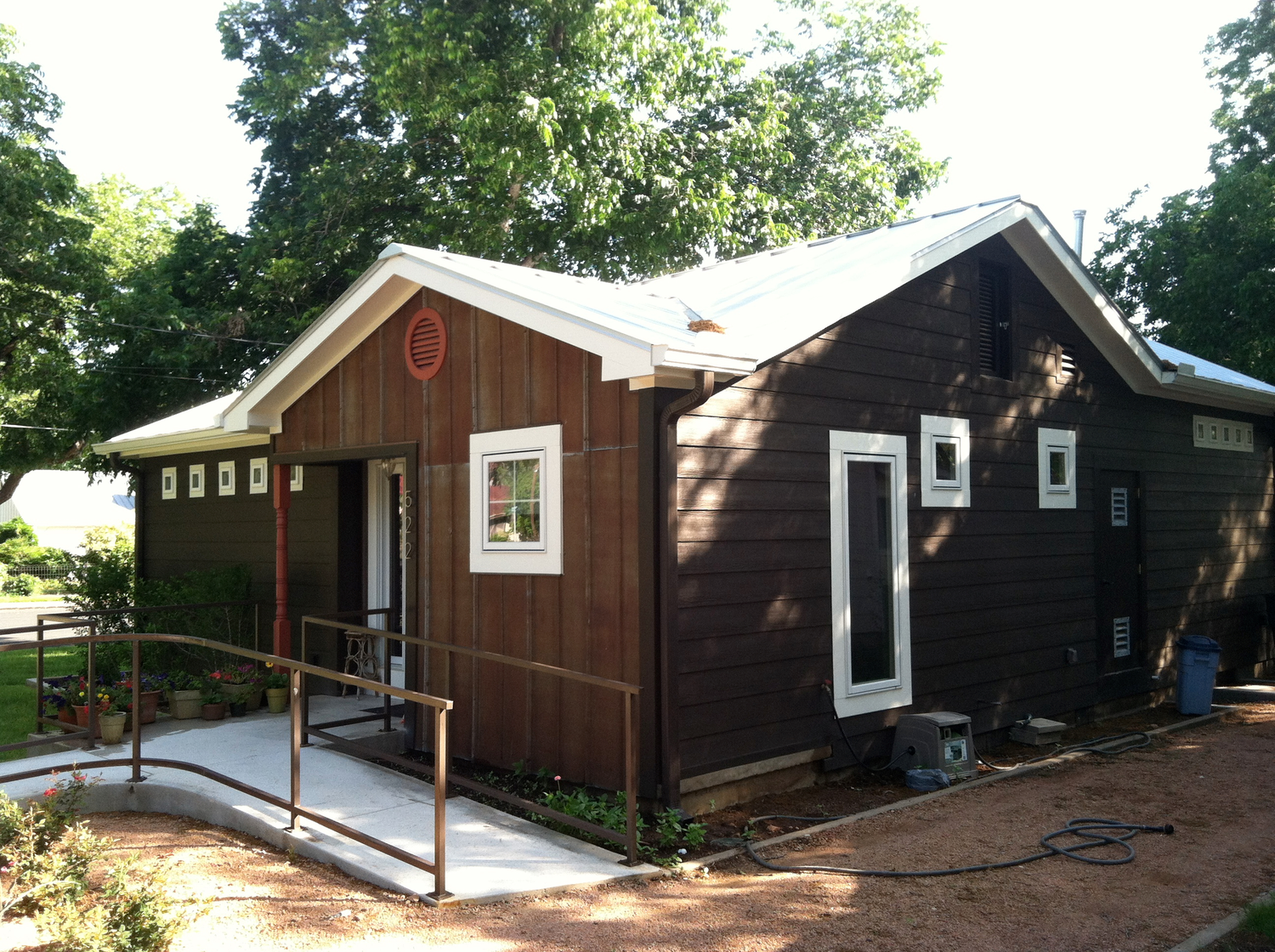 Small brown ranch house with white trim and wheelchair accessible ramp sits on grassy lot with mature trees.