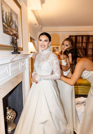 Bride getting ready in white wedding dress with helpers assisting in final preparations before ceremony.