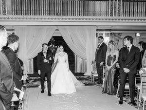 A wedding ceremony procession in a grand hall with white draping and guests looking on.