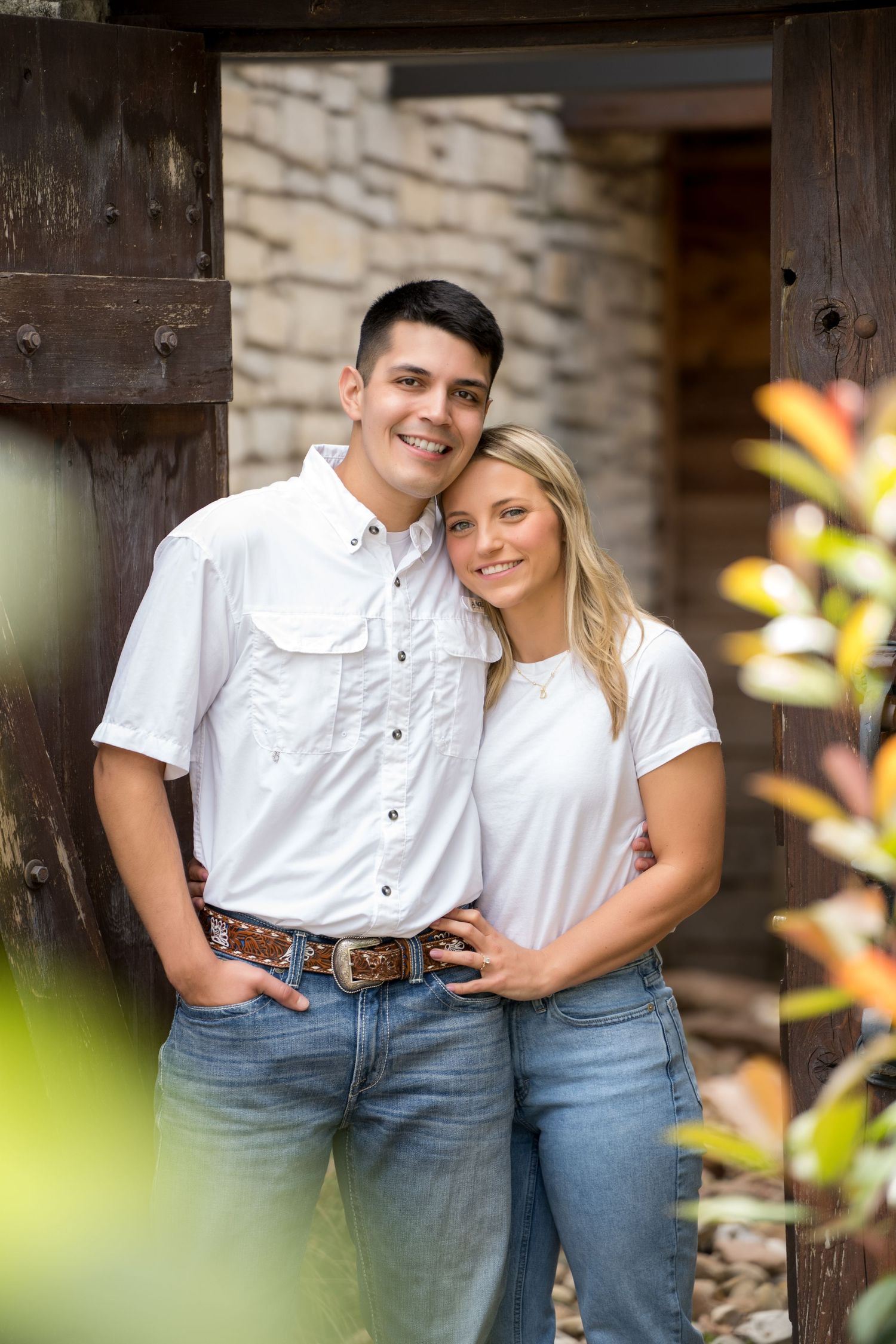 A smiling young couple in white shirts and jeans pose together in front of rustic wooden doors and stone wall.