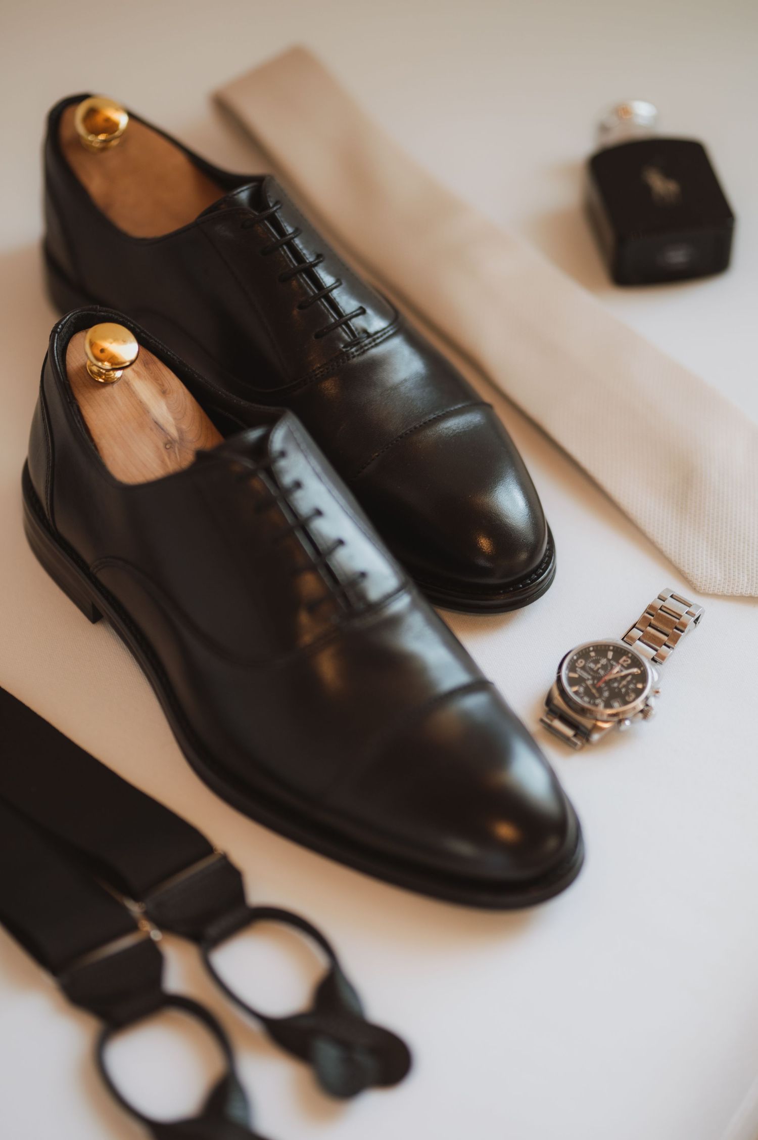 Black leather dress shoes and accessories arranged on white surface.