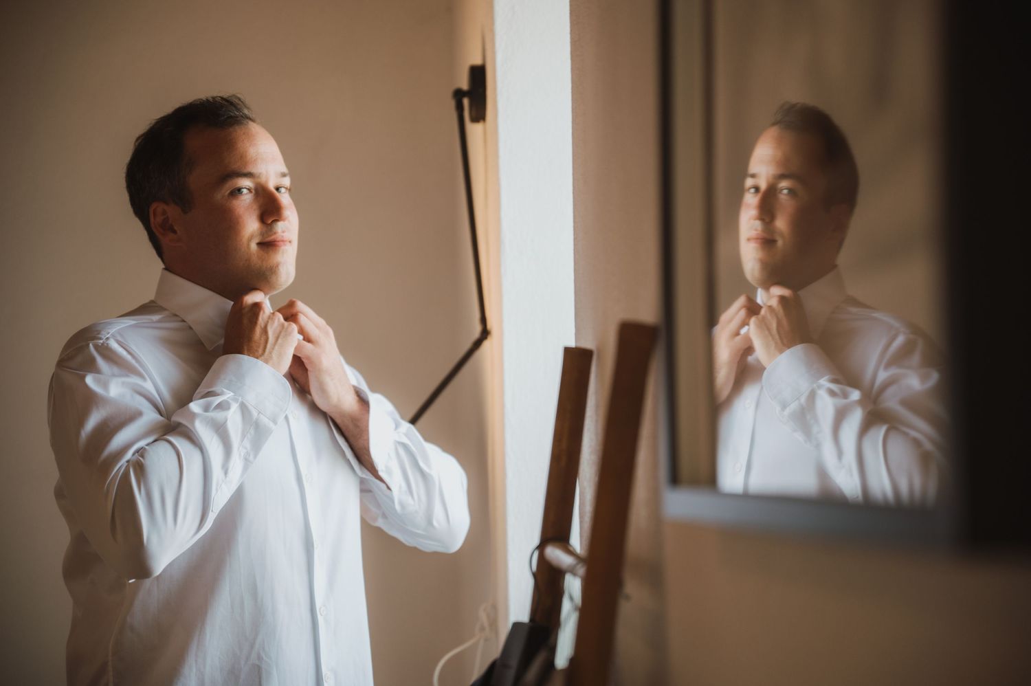 Groom prepares for wedding day in soft window light while adjusting his tie.