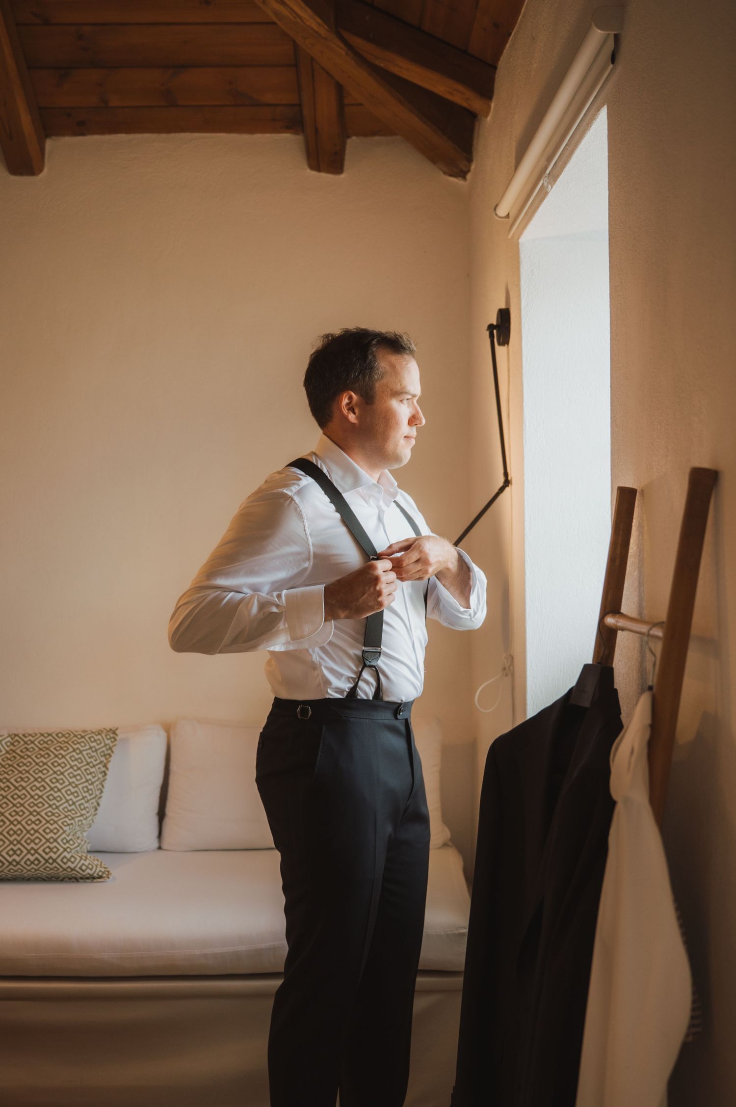 A person adjusts suspenders while getting dressed near a window.