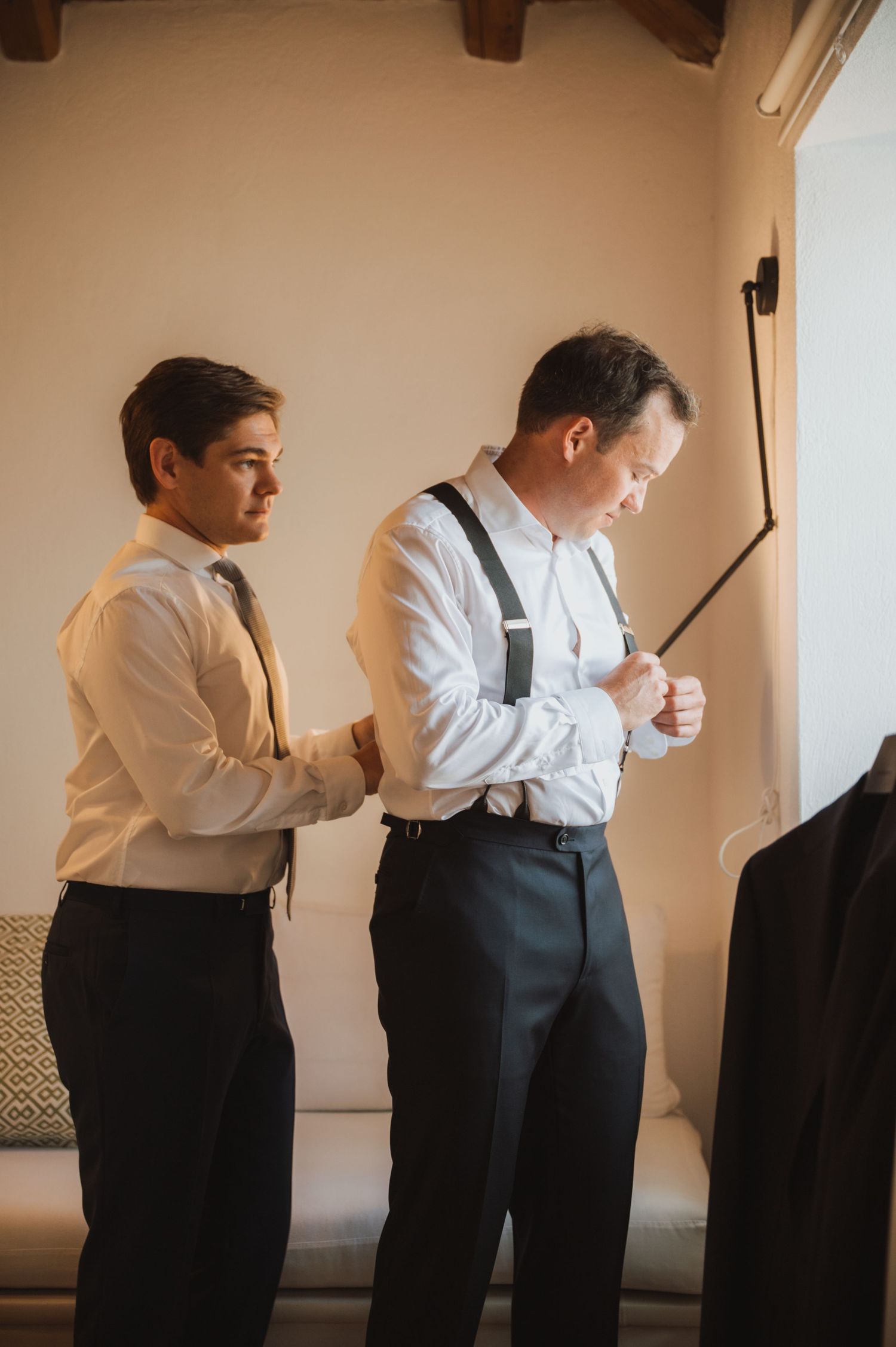 Two men in formal attire prepare for an event in a minimalist room with wooden beams.