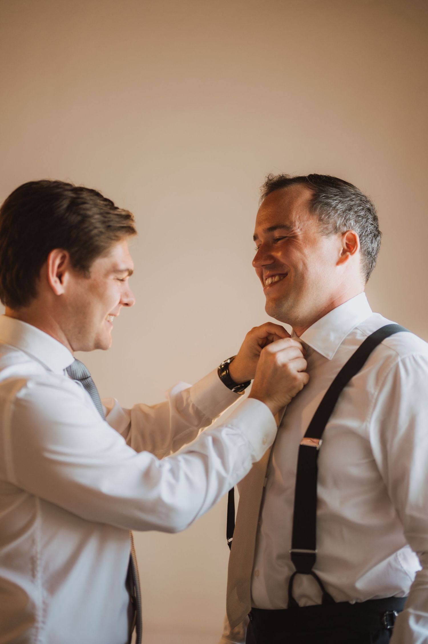 Two people in white shirts and suspenders share a joyful moment while getting ready.