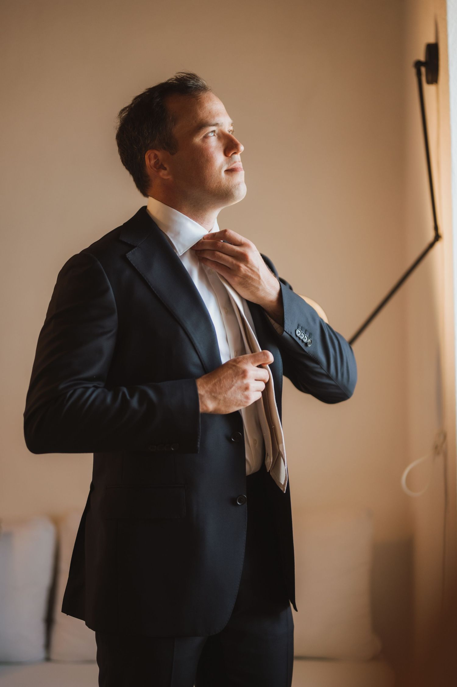 A person in a dark suit adjusts their tie while standing in natural light.