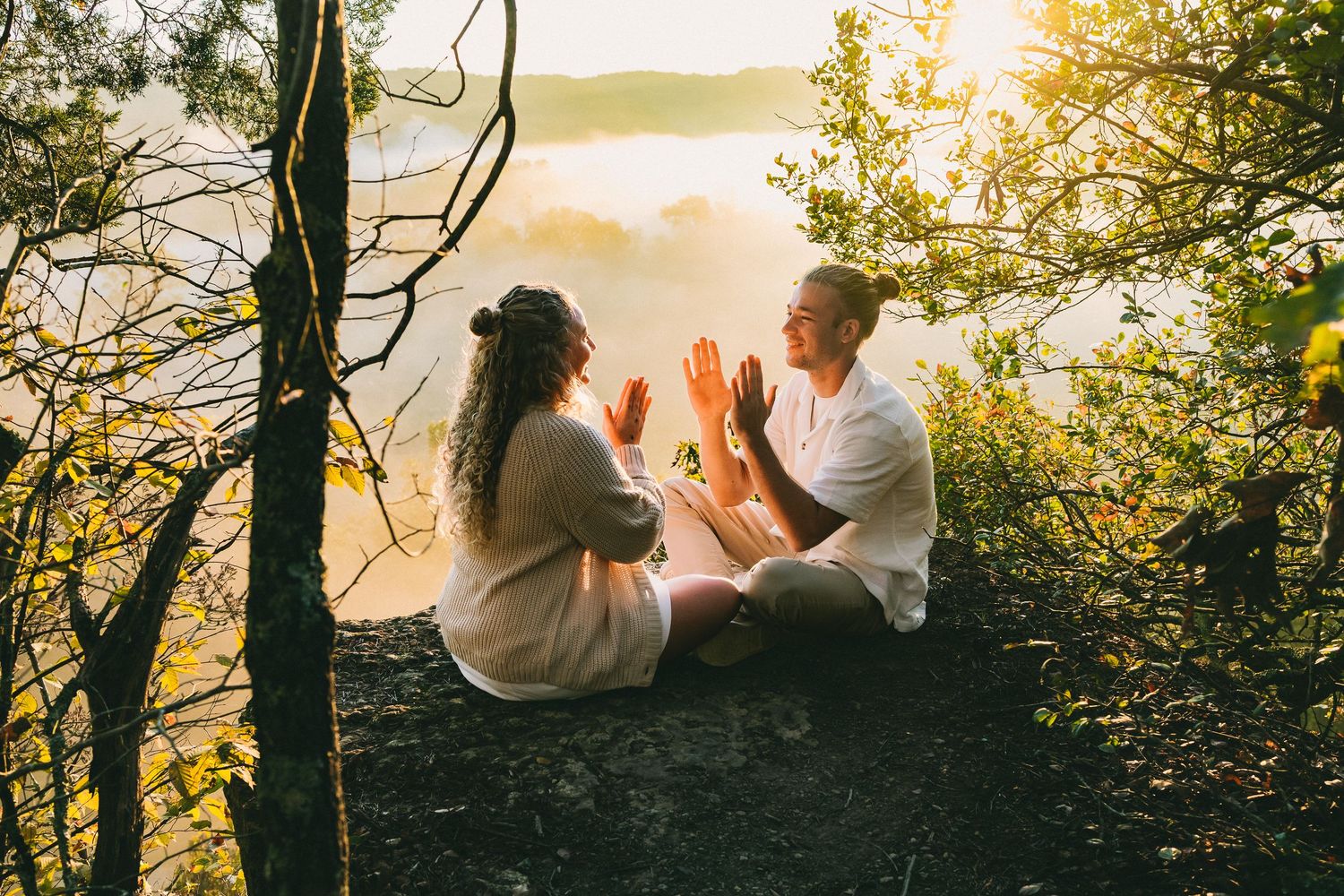 A couple sits together on a cliff edge enjoying a scenic sunset through tree branches at golden hour.