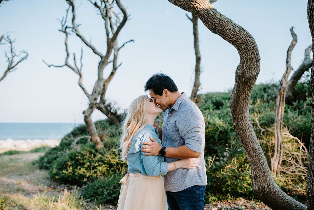 A romantic couple embraces under twisted trees near a coastal beach at sunset, creating a dreamy seaside atmosphere.