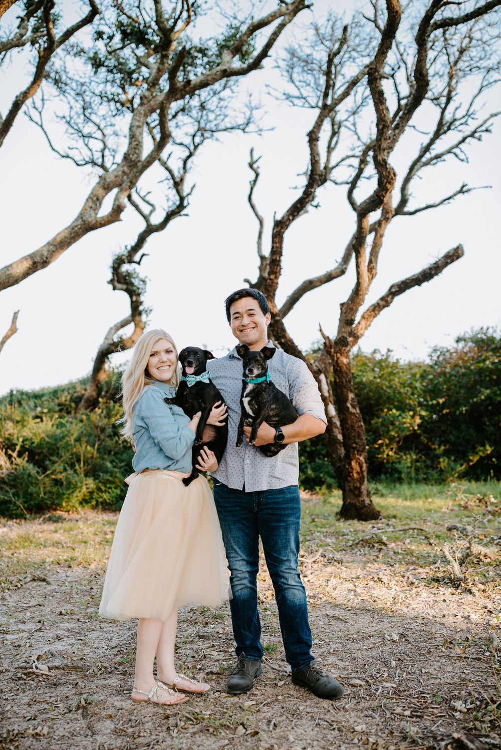 A couple stands together with their black dog on a scenic hilltop path surrounded by twisted trees in nature.
