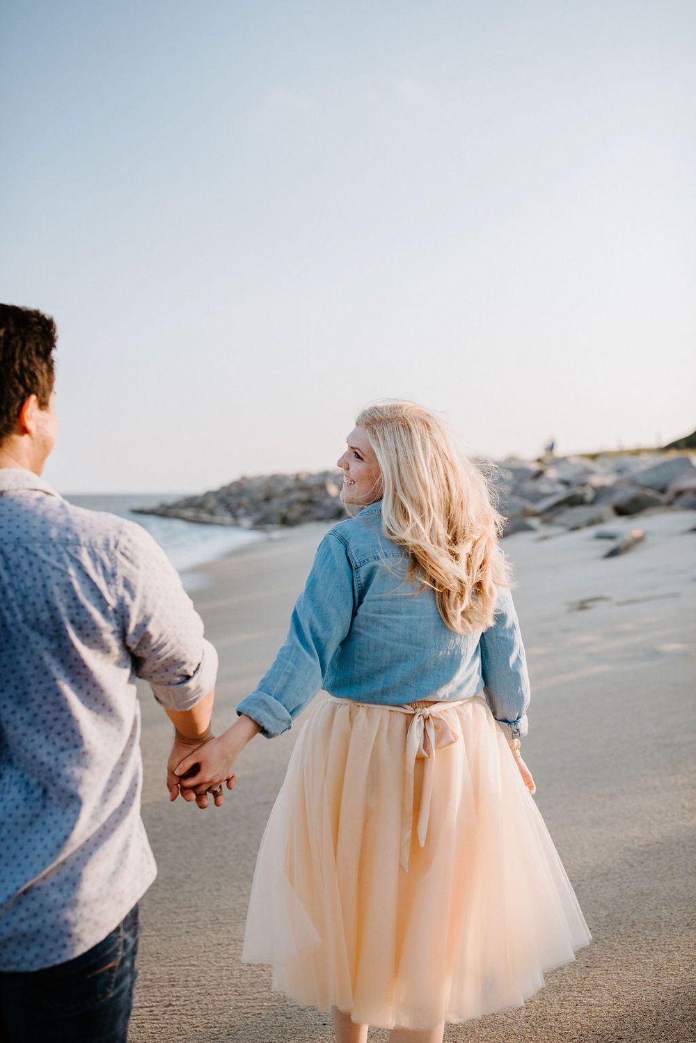 A couple holding hands walks along a sandy beach, with the woman wearing a flowy peach tulle skirt and denim top.