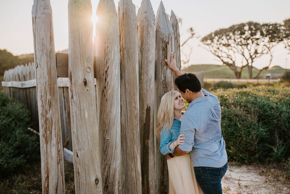 Couple sharing romantic moments against rustic wooden fence posts during golden hour sunset.