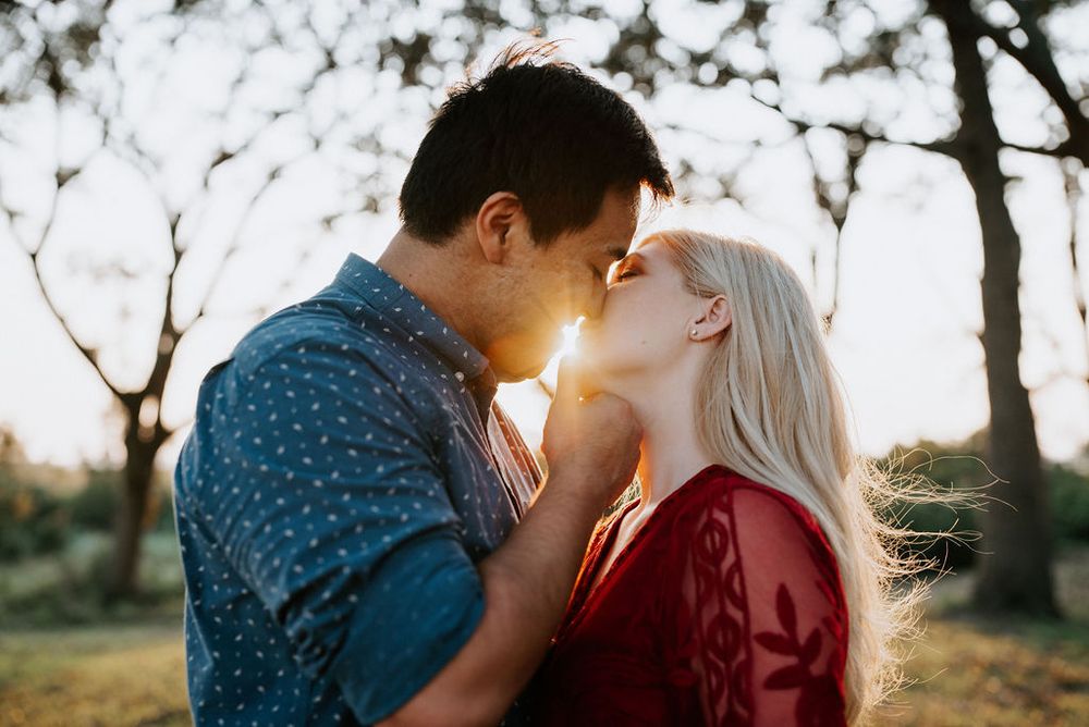 A romantic couple shares a tender kiss in a sunlit outdoor setting wearing blue and burgundy clothing.