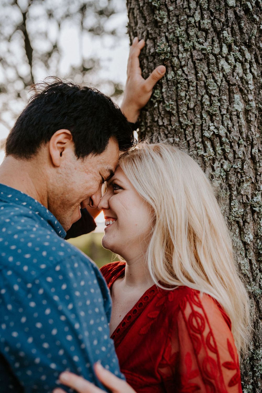 A romantic couple shares an intimate moment while standing close together against a tree trunk background.