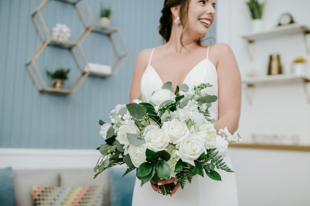 A beautiful white wedding bouquet with roses and greenery is held against a light blue wall with floating shelves.