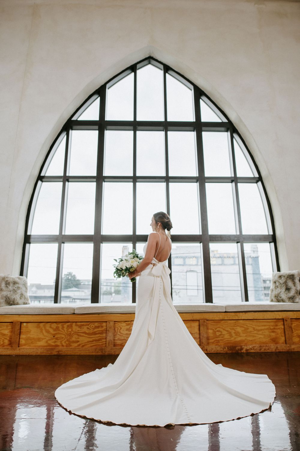 Elegant white wedding dress with long train silhouetted against dramatic arched cathedral window.