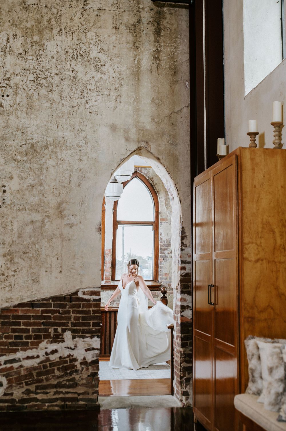 A bride in a white wedding dress poses in a rustic venue with exposed brick walls and wooden doors.