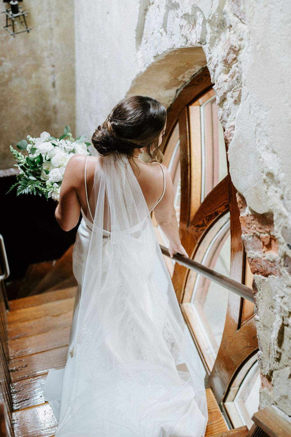 A white wedding veil flows gracefully down a rustic wooden staircase in an old building with textured walls.