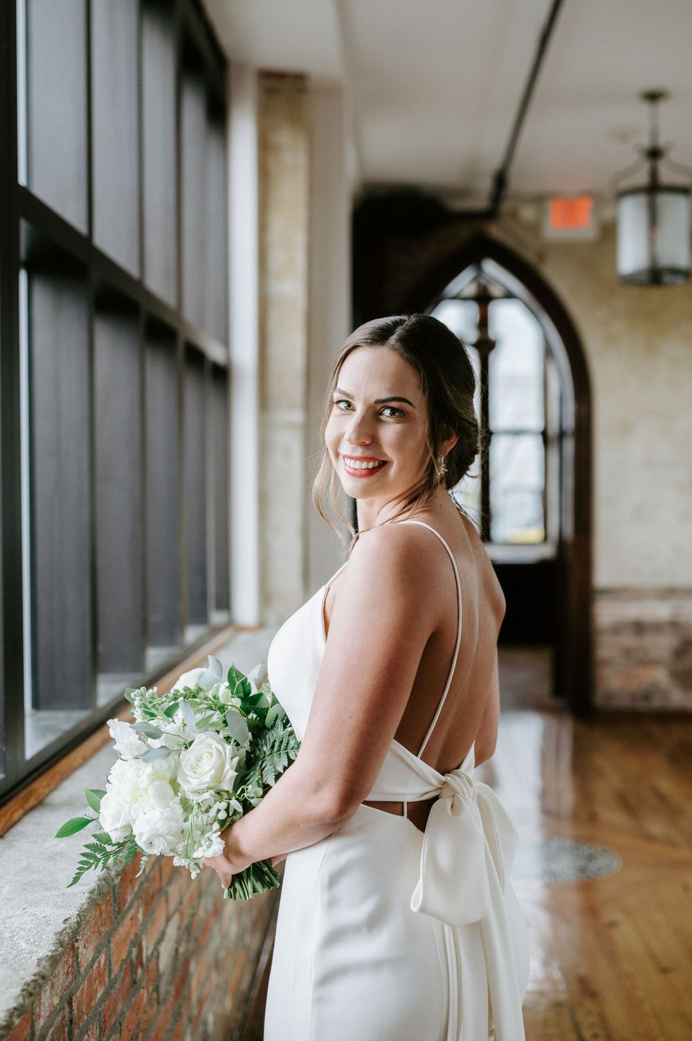 A bride in a white wedding dress poses elegantly in a bright indoor venue with wooden floors and large windows.