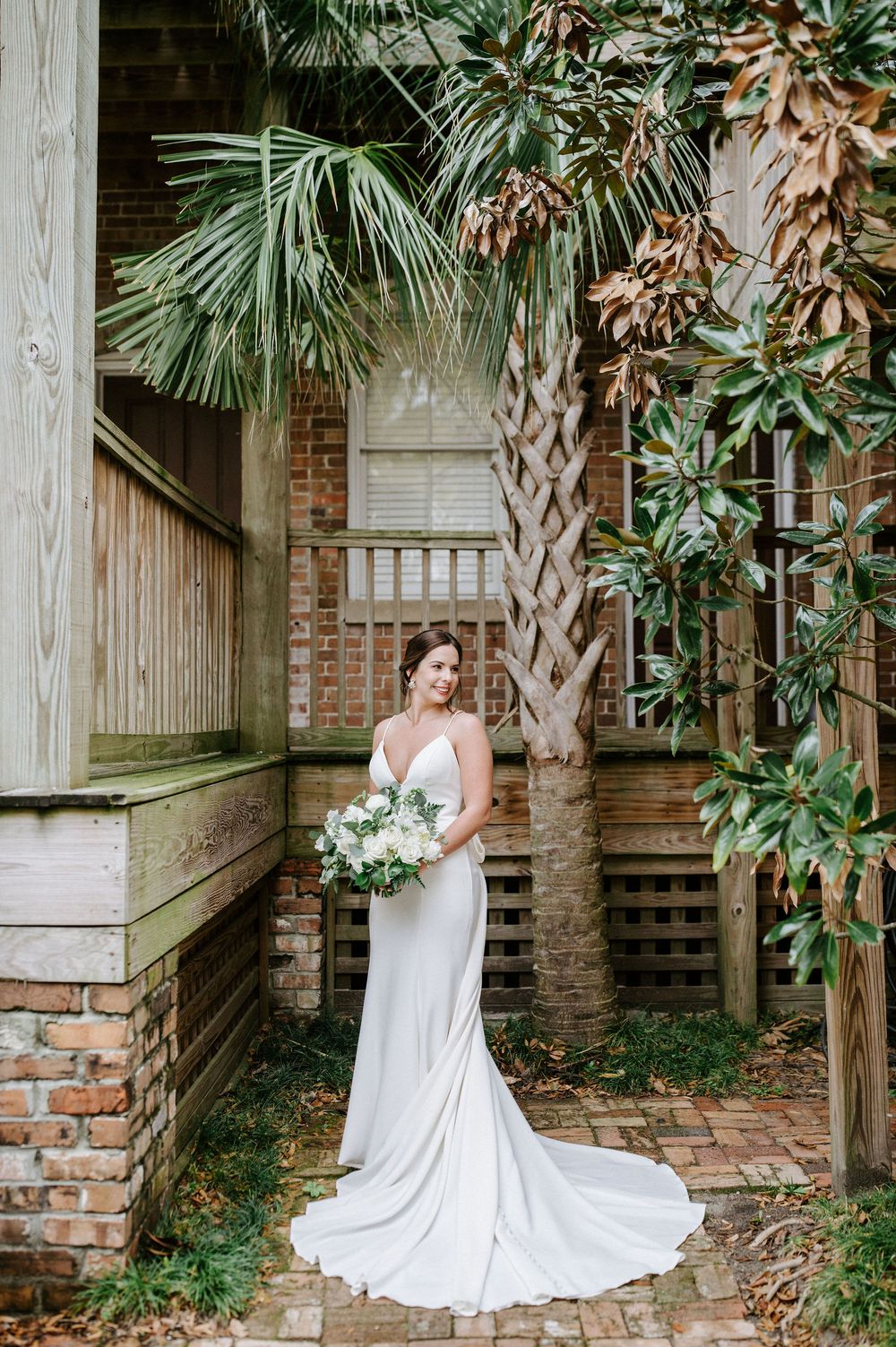 A bride in a flowing white wedding dress poses elegantly against a rustic brick and wood backdrop with tropical palm trees.