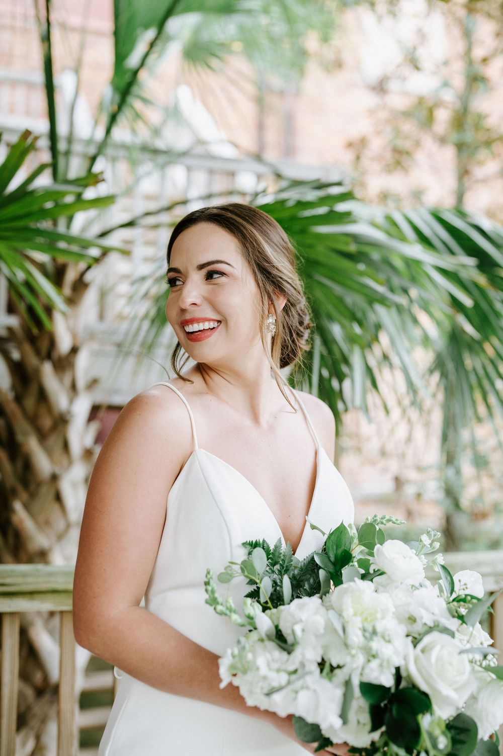 Bride in white wedding dress holds elegant white floral bouquet while standing near tropical palm trees.