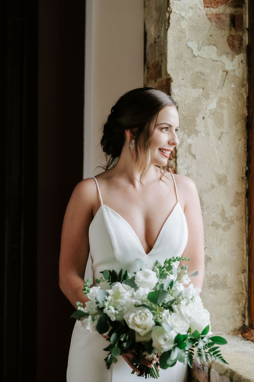 A bride in a white spaghetti strap wedding dress holds an elegant bouquet of white roses and greenery by a rustic window.