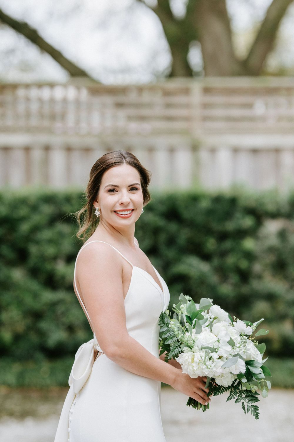 A bride in a classic white wedding dress holds a lush bouquet of white flowers and greenery in an outdoor garden setting.
