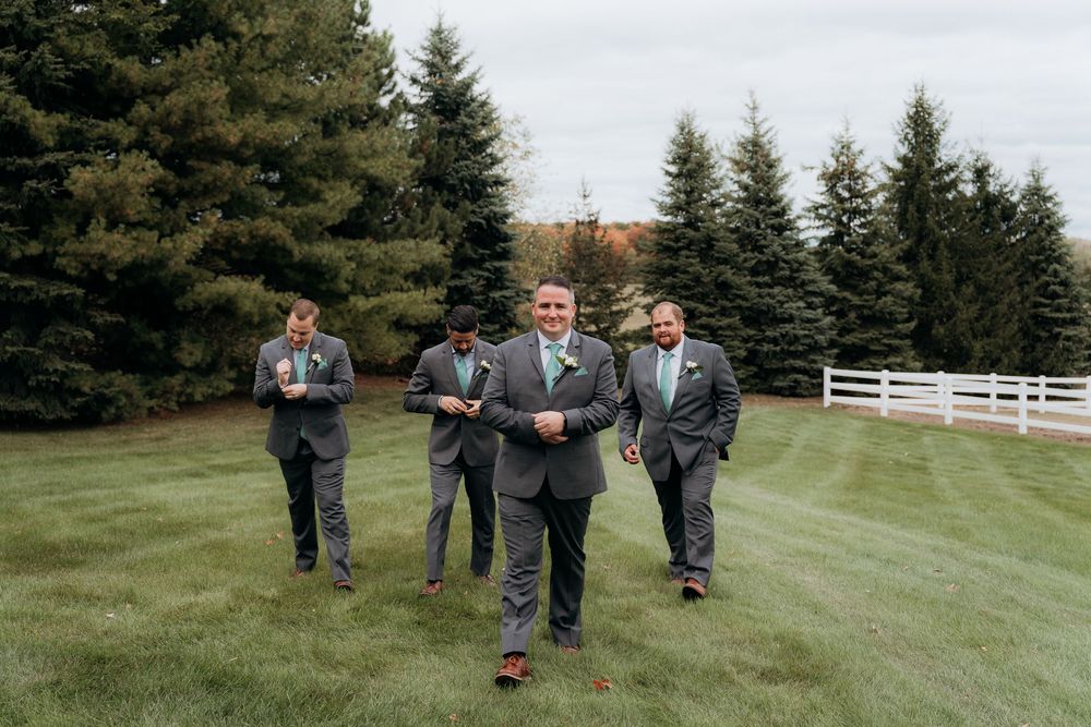The groom and groomsmen walk and pose together on a grassy lawn with evergreen trees and white fencing in the background. Garvey Farm.