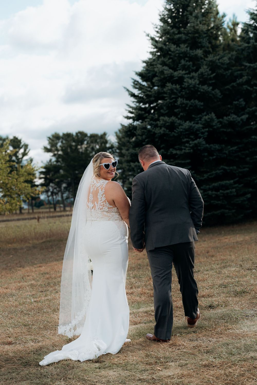 A bride and groom walk away together across a grassy hillside with evergreen trees in the background. Garvey Family Wedding Farm TC Michigan