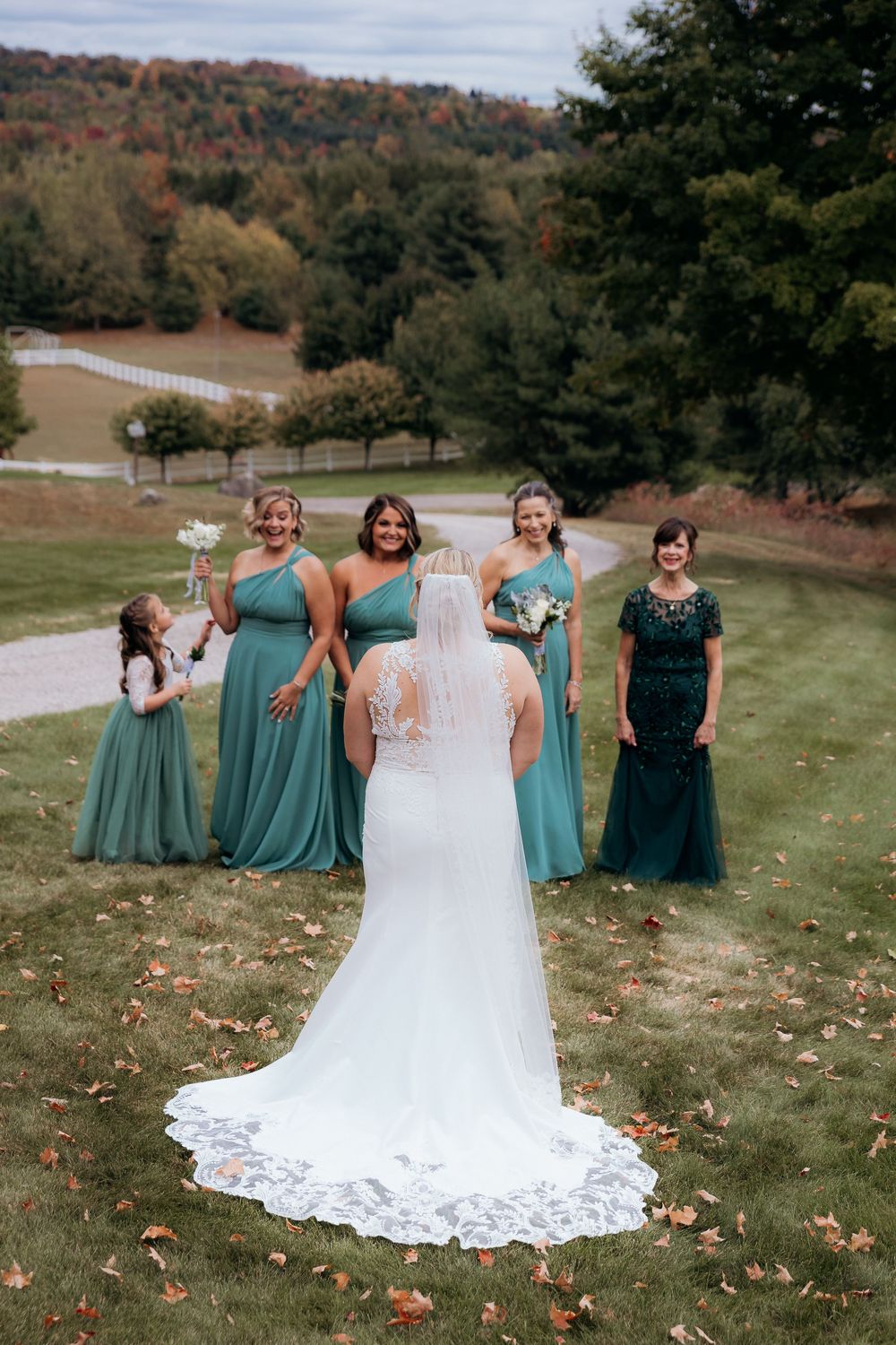 Bridesmaids in teal dresses gather around the bride's lace wedding gown on a grassy hillside. First Look for bridesmaids.