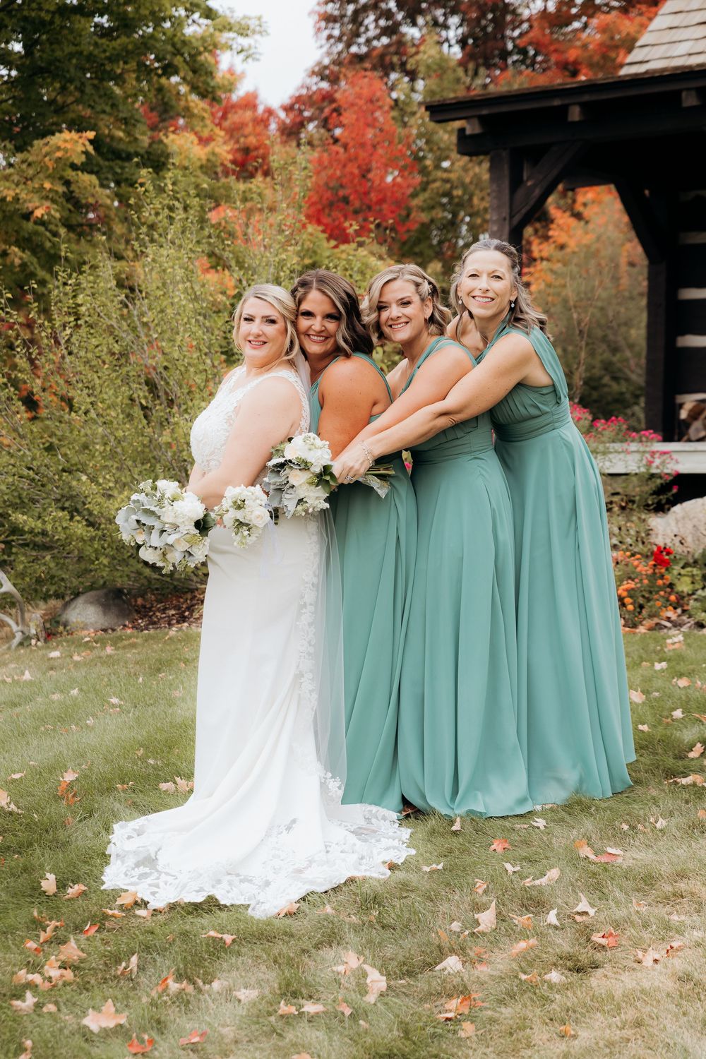 Wedding party in green teal dresses poses together on an autumn lawn with colorful fall foliage. Garvey family wedding barn traverse city MI