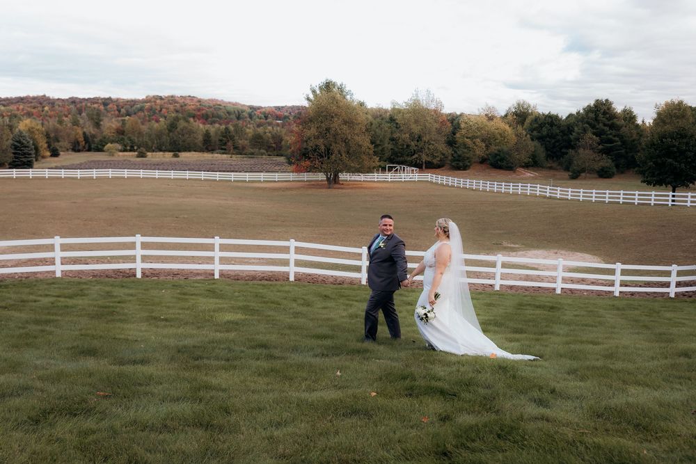 Wedding couple posing along a white fence on rolling countryside grounds.