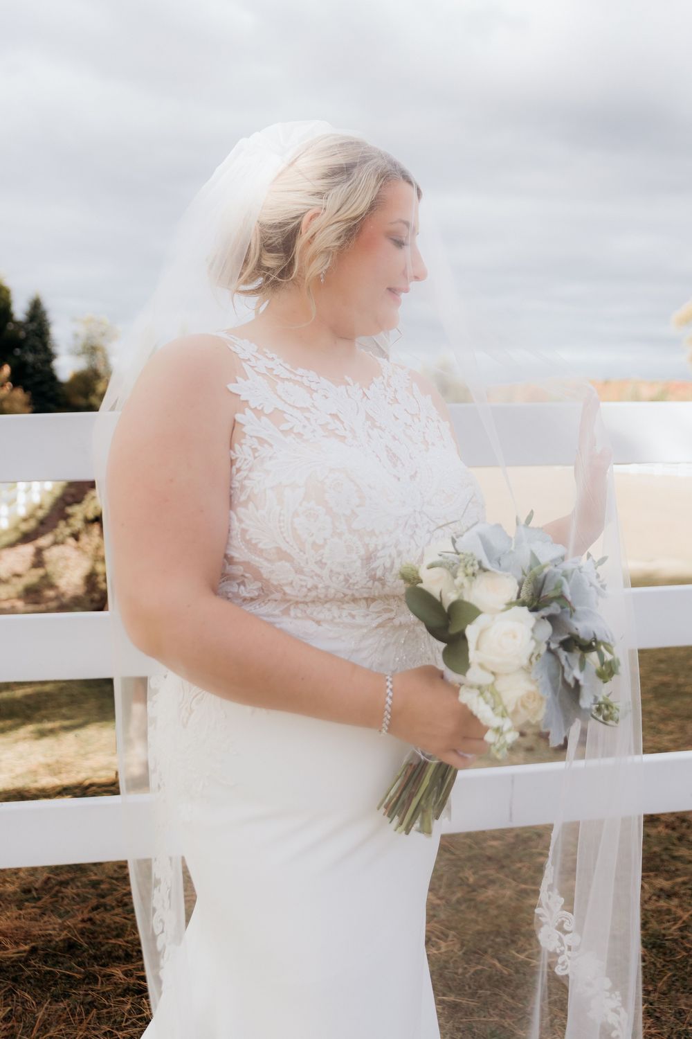Bride holding an elegant white bouquet against a white fence on a sunny autumn day. Bridal Portraits. Garvey wedding barn Traverse City MI